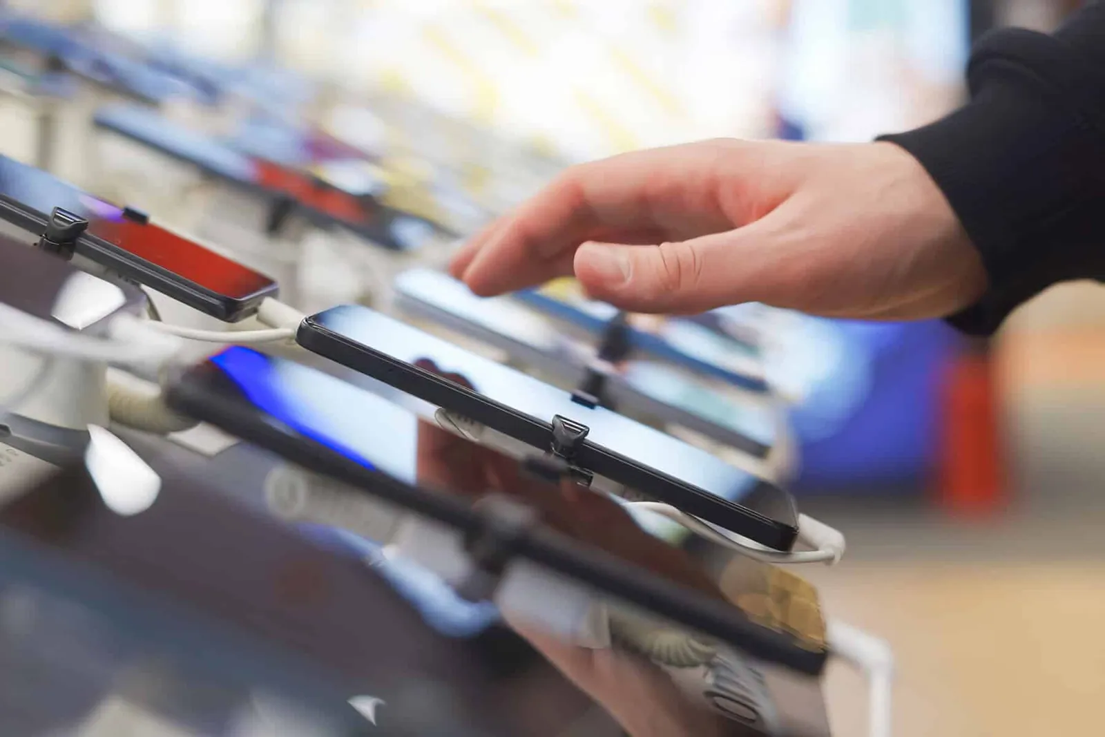 Hand reaching toward smartphones on security-tethered display stands in an electronics store.