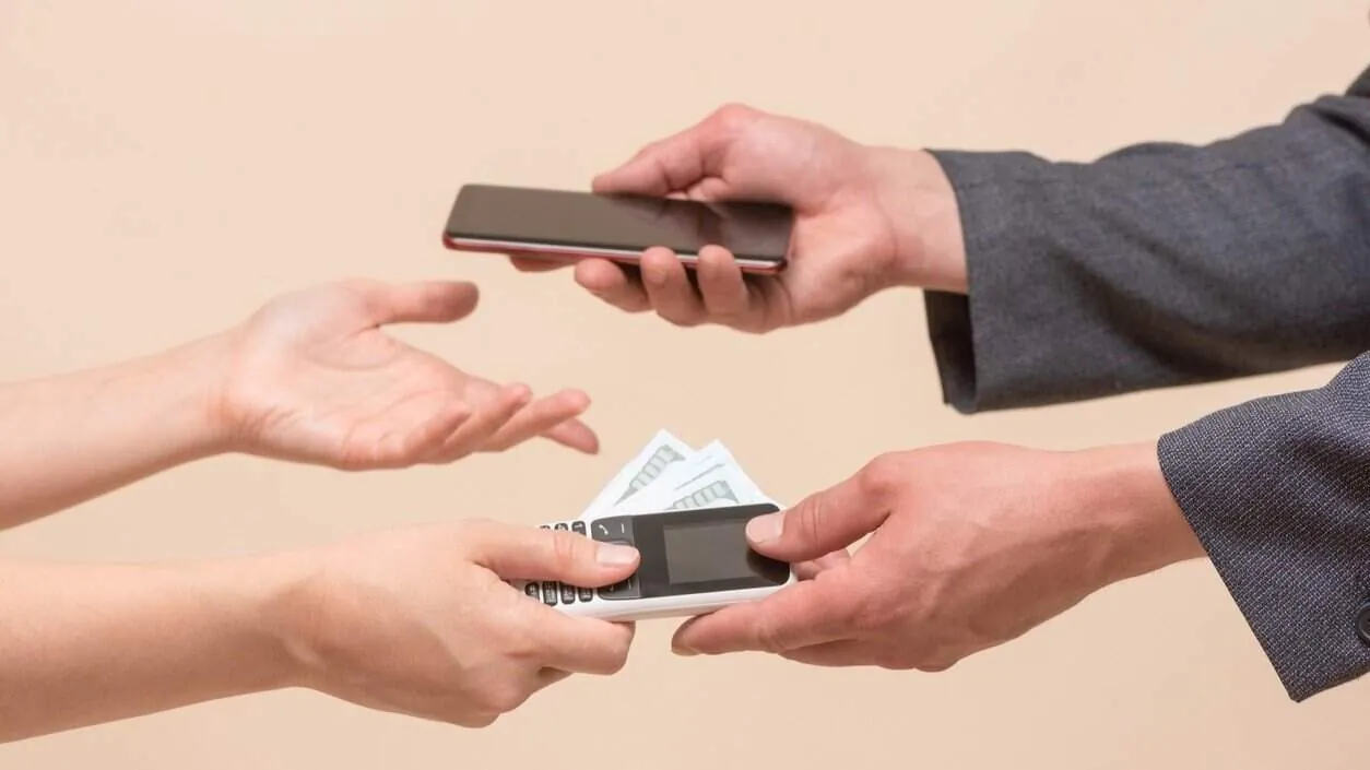 Hands exchange a smartphone for an old keypad phone and cash between two people.