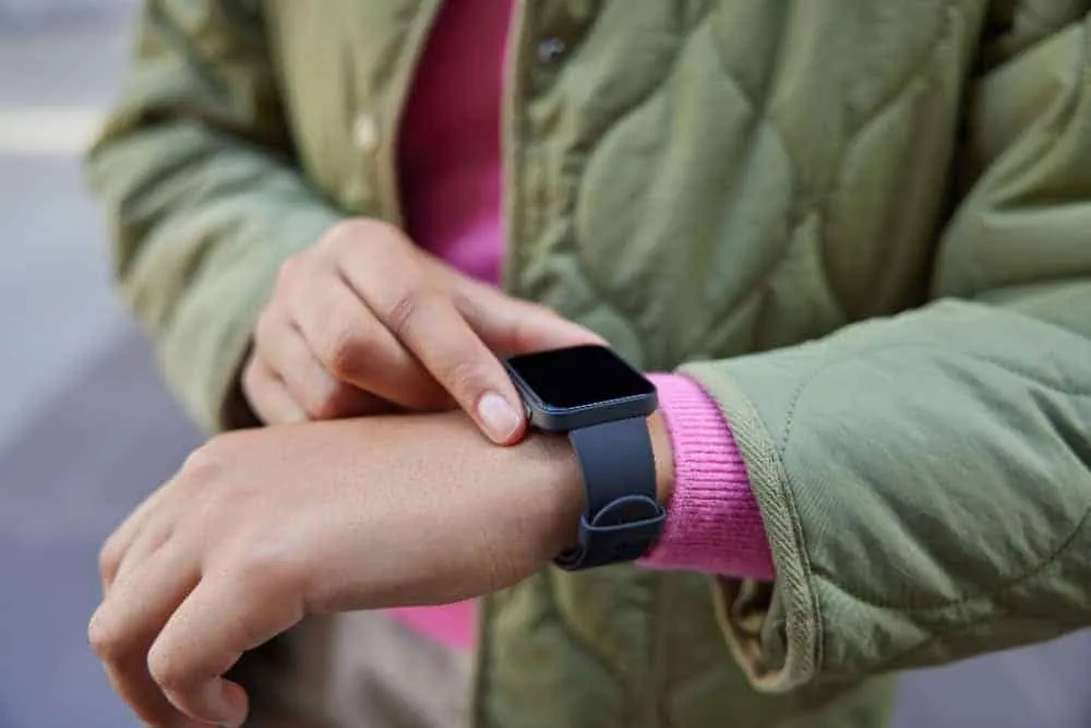 Person in a green quilted jacket touching a square black smartwatch on their wrist.