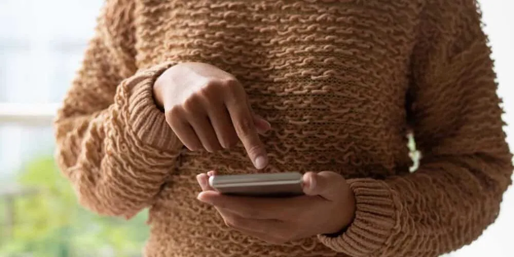 Person in a brown knit sweater tapping the screen of a smartphone held in one hand.