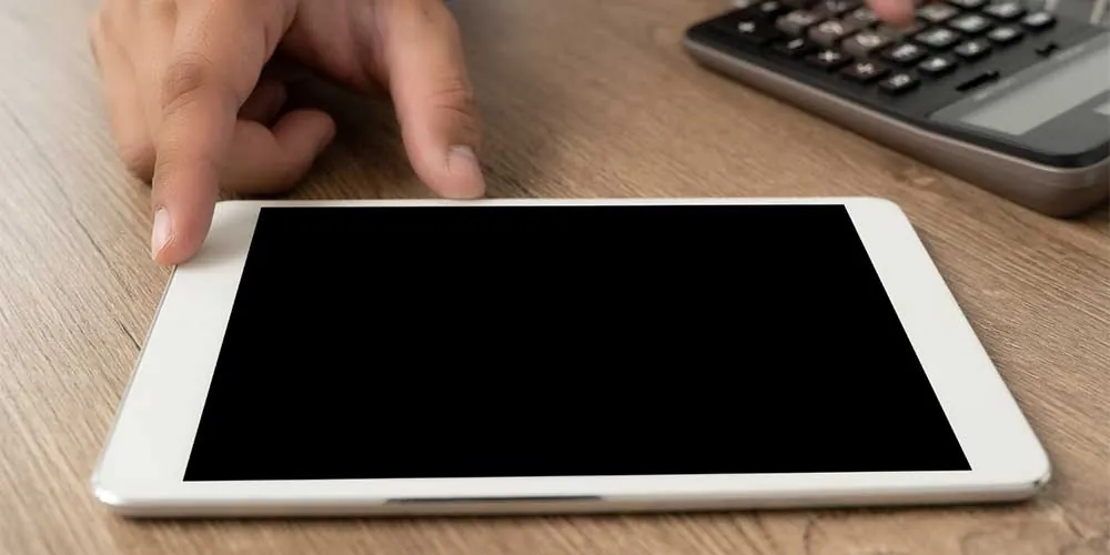 Fingers touch the edge of a white tablet with a blank screen on a wooden desk next to a calculator.