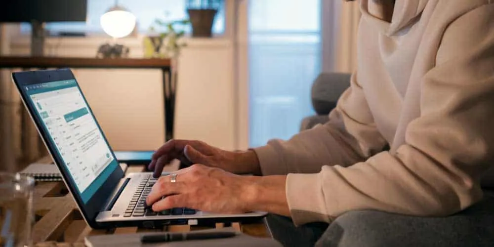 Person in a cream hoodie typing on a laptop at a low table in a dimly lit living room.