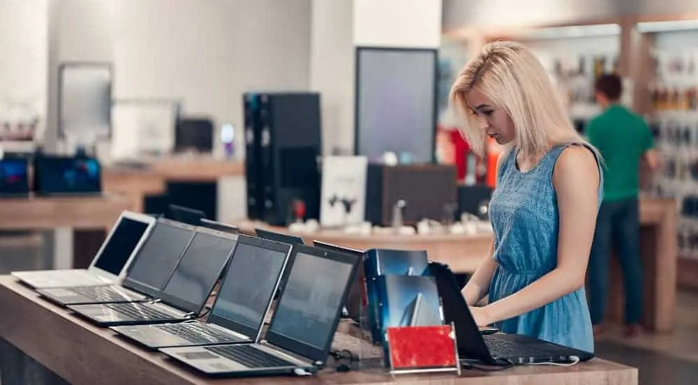 Woman in a blue dress browsing a row of laptops on a display table in an electronics store.